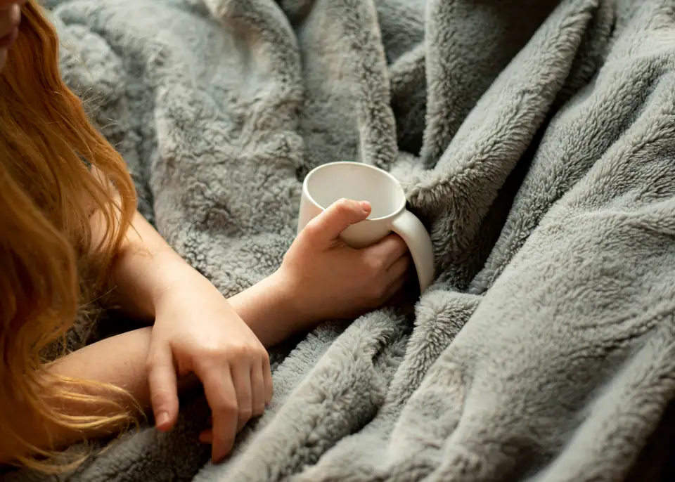 Hands holding a white mug under a thick grey fleece blanket