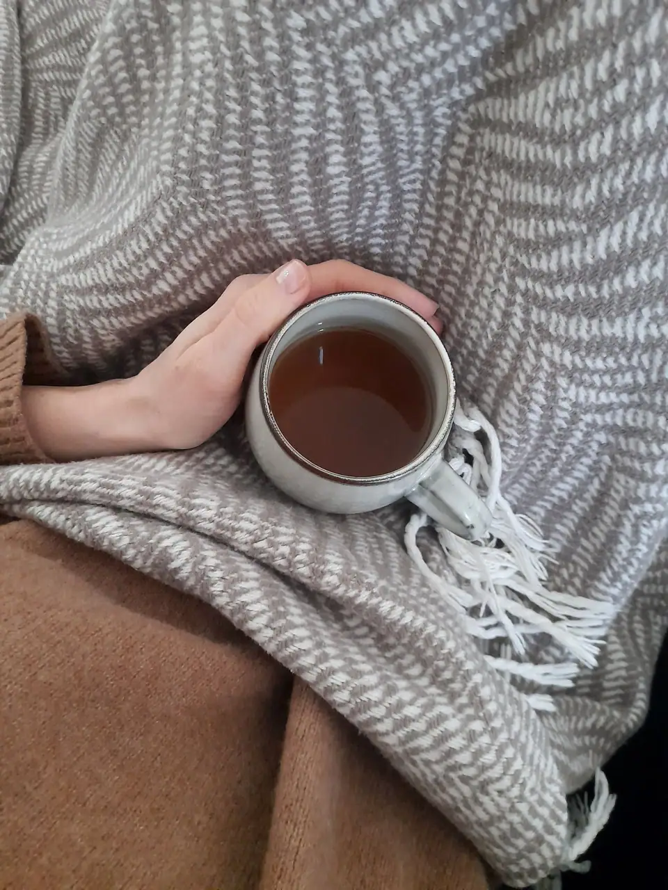 A hand holding a mug of tea on a gray woven throw