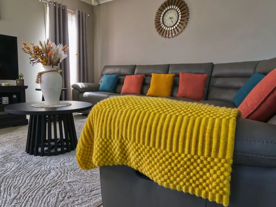 A mustard yellow chunky knit throw over the arm of a gray sofa with colorful cushions